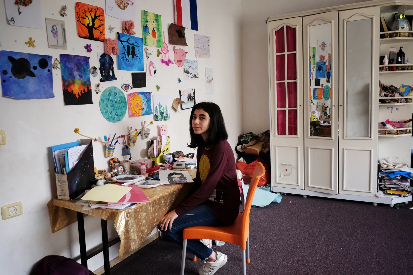 Teen sits at her desk by a wall filled with her drawings at her home. 