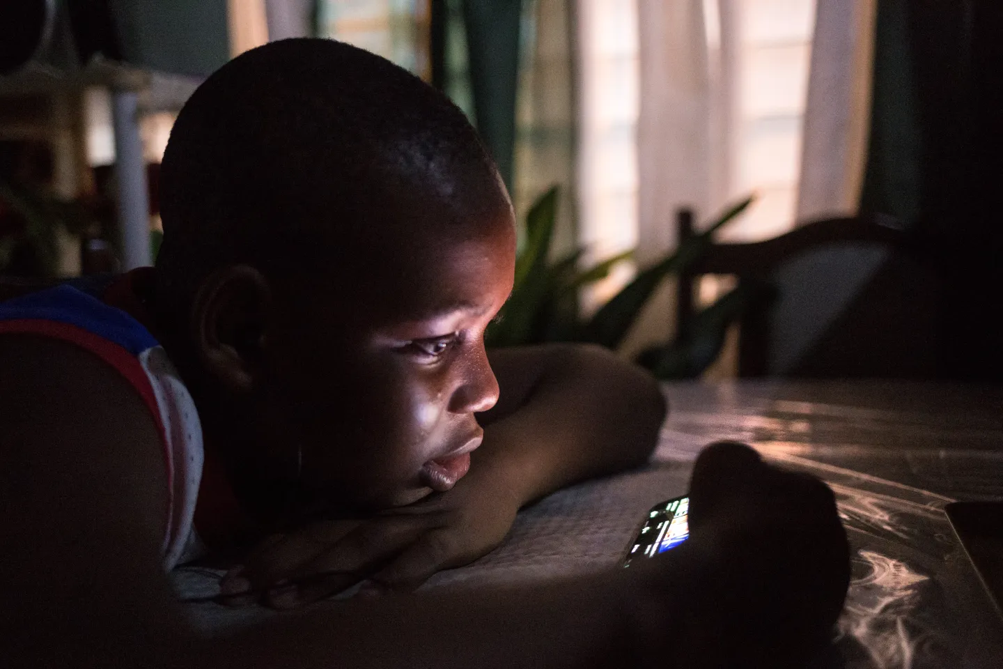 A teenager plays a game on a mobile phone while lying in bed at home