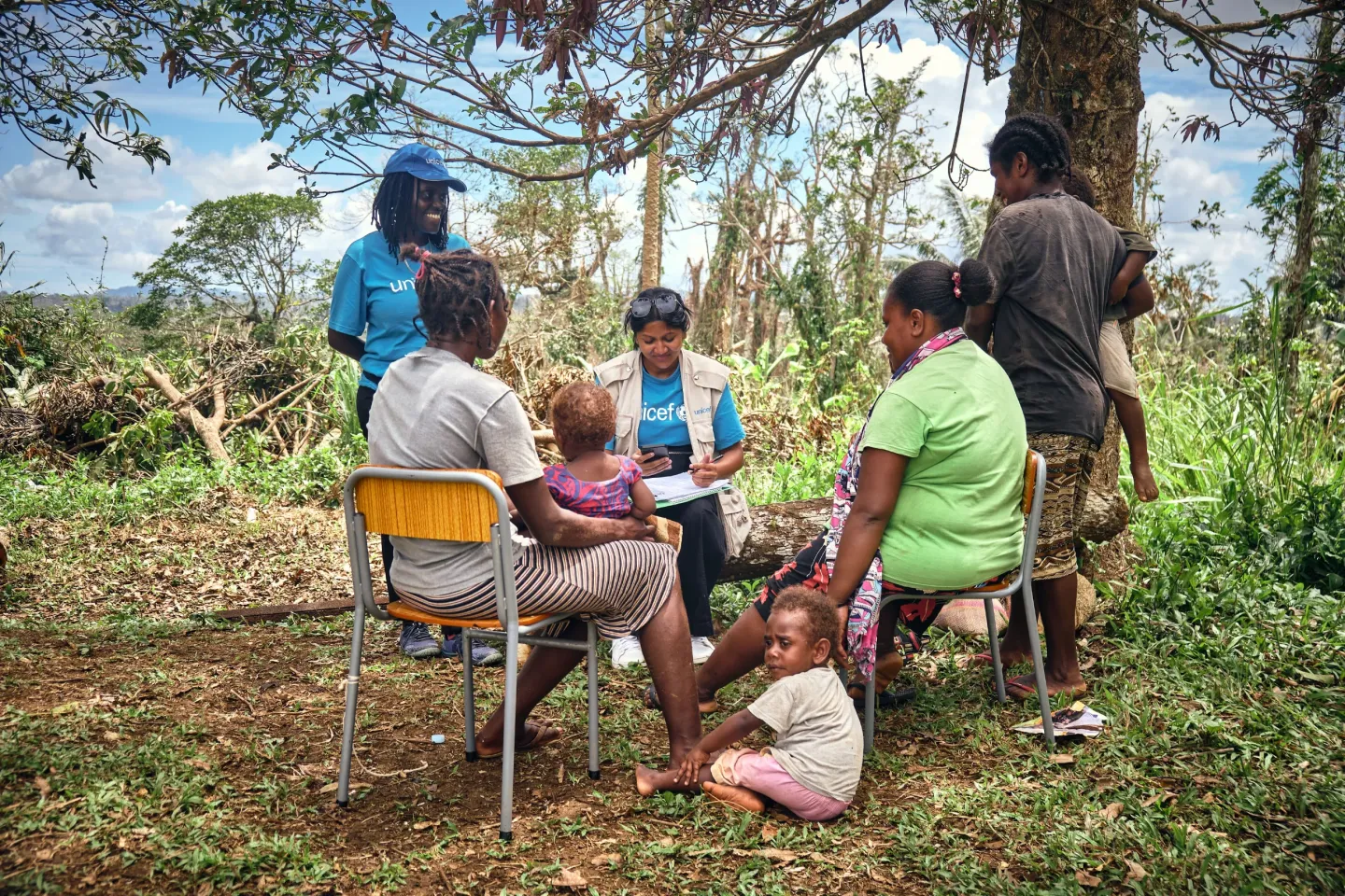 UNICEF staff speaks with a small group of mothers and babies, while sitting in chairs under a tree