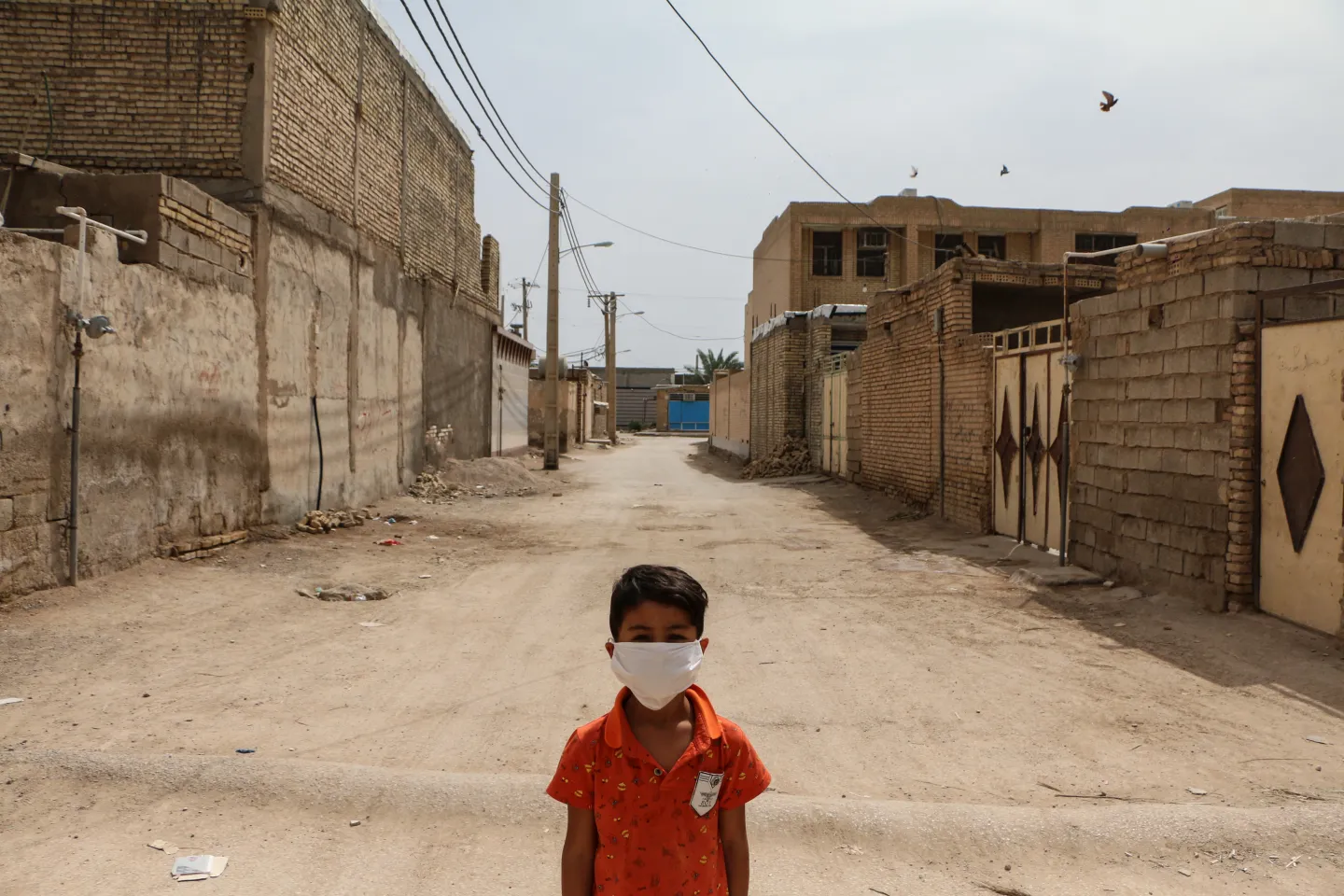 A child wearing a medical mask standing in the middle of a dirt road the suburbs of Ahvaz, Iran