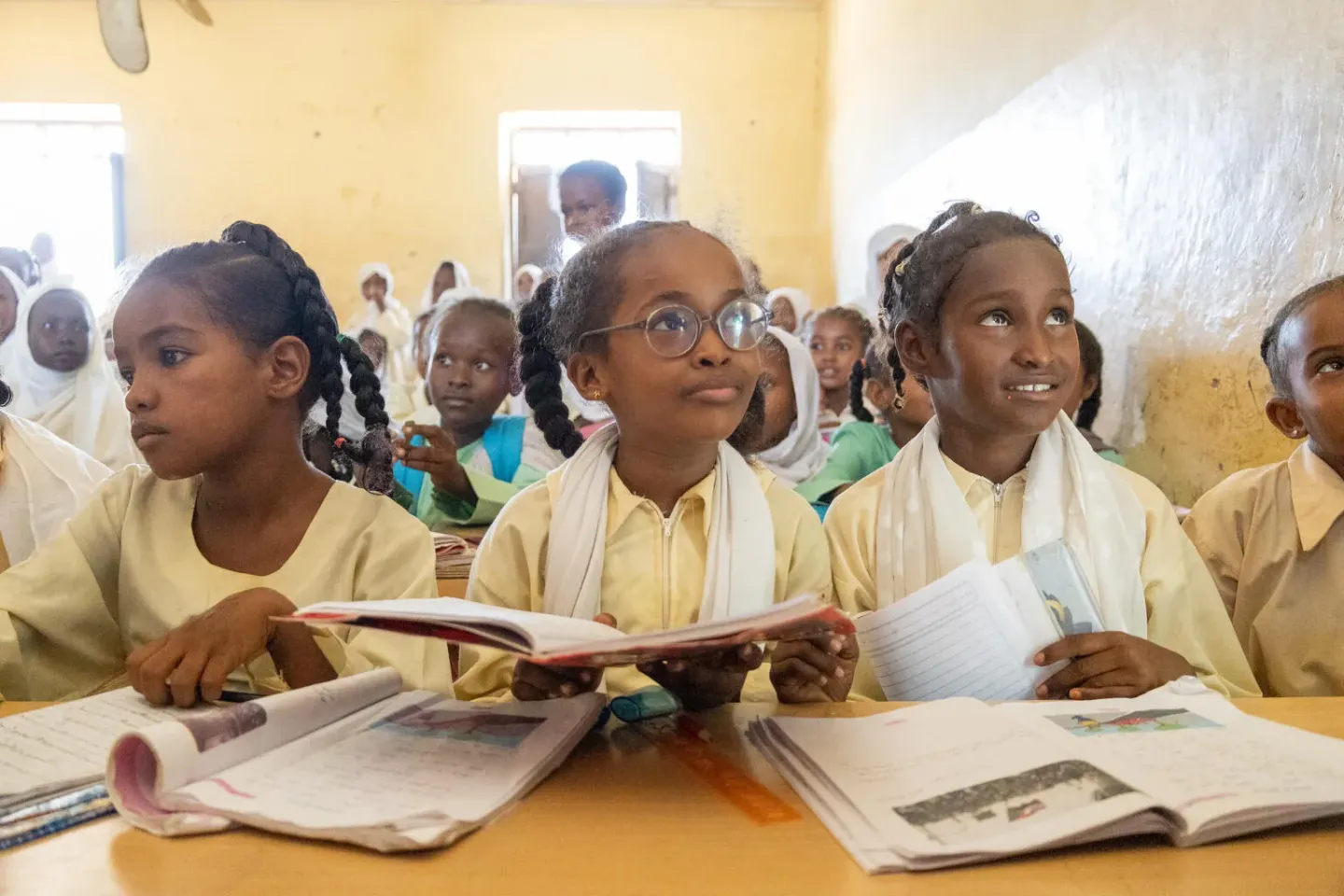 Children seated in a classroom wearing yellow and white uniforms with books open in front of them. Their gaze is towards an object behind the camera, possibly a teacher or a board, as they are paying attention to the lesson.