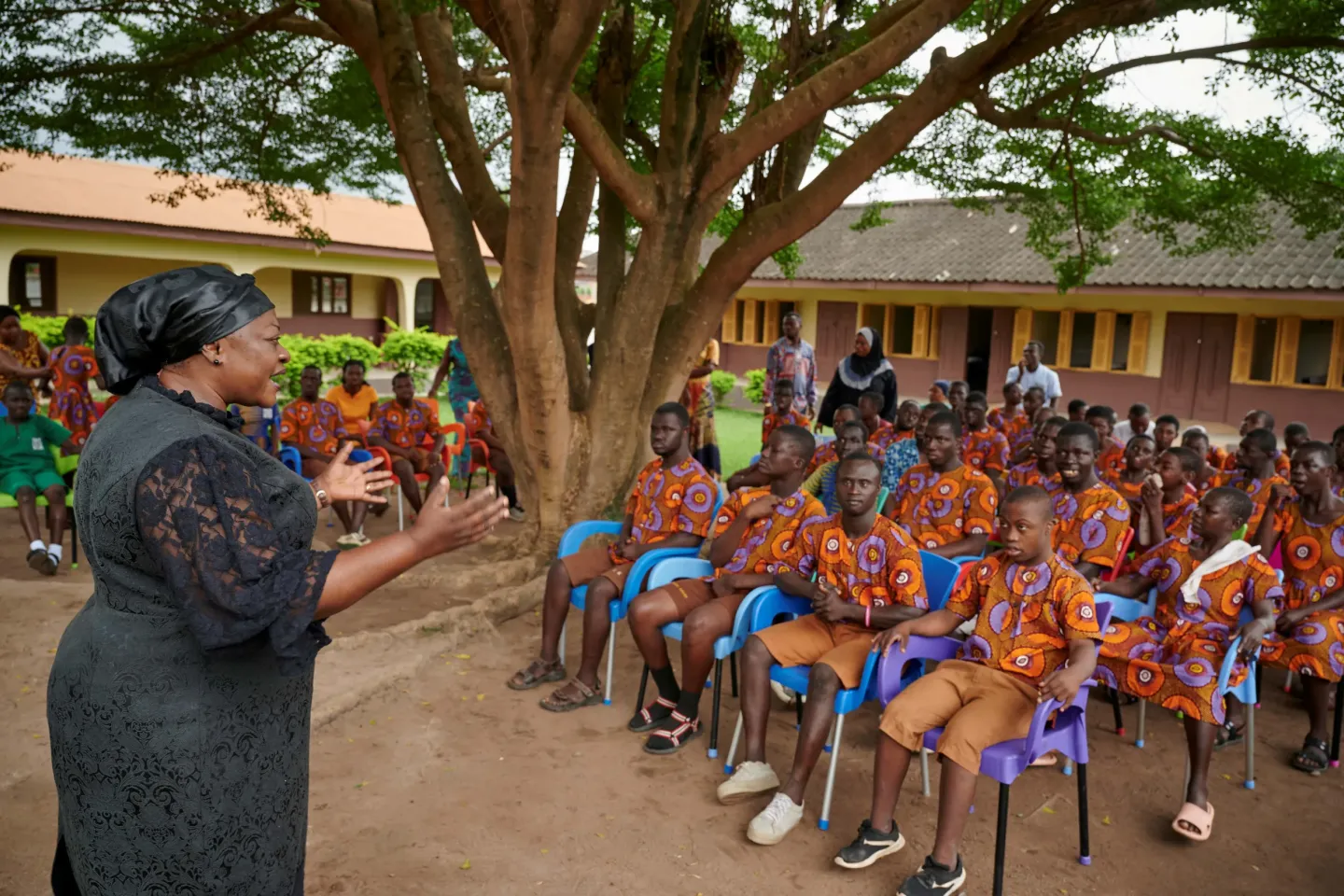 A female professor stands before a group of students who are sitting in front her, listening to her speak, in a patio under a tree and with a building that looks like a school in the background.