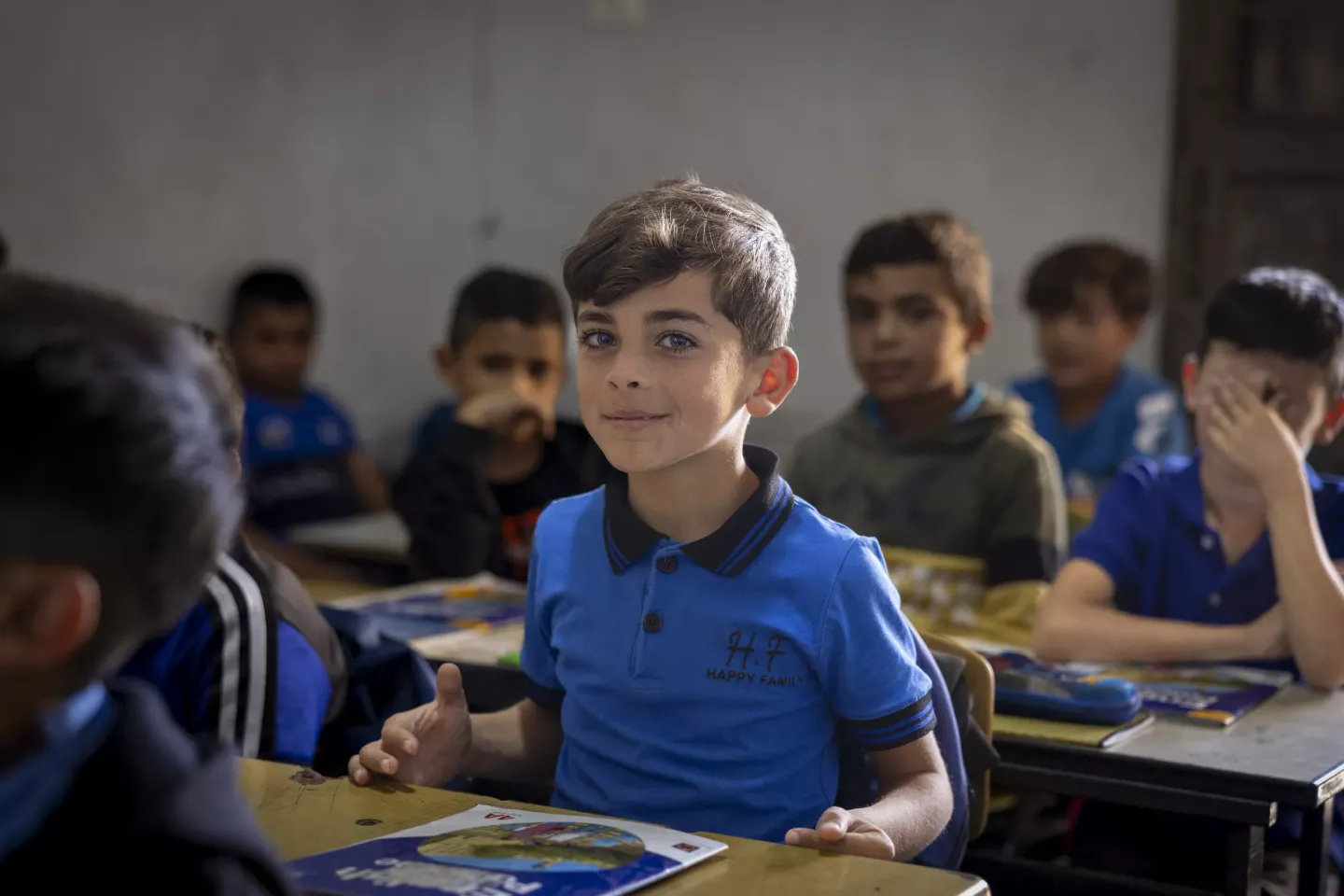 A boy sitting at his desk in a classroom. He is looking at the camera and smiling