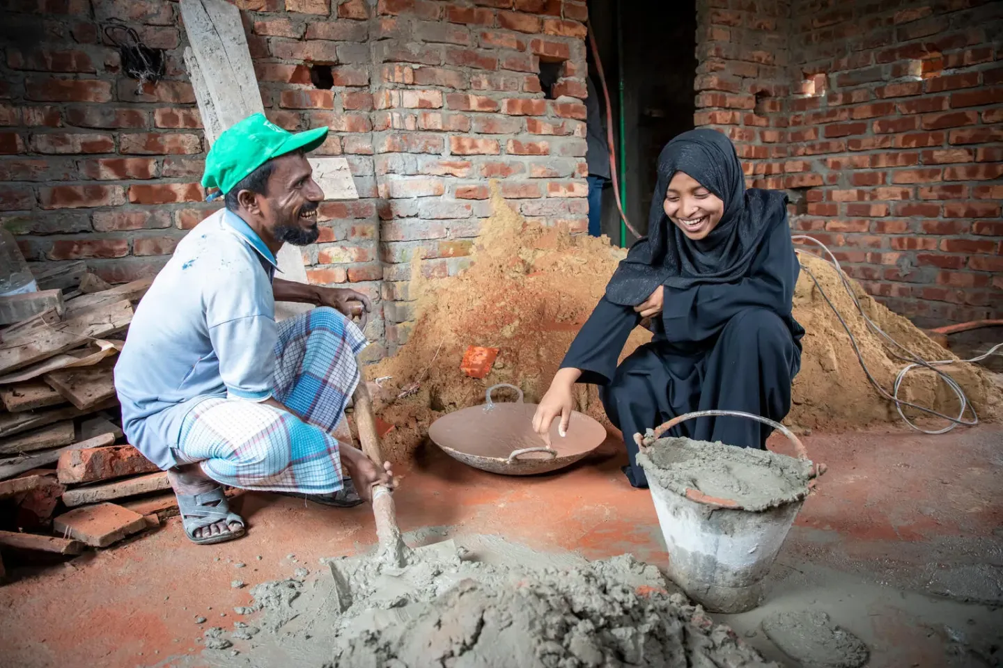Two people working at a contruction site, one shoveling cement, the other giving instructions 