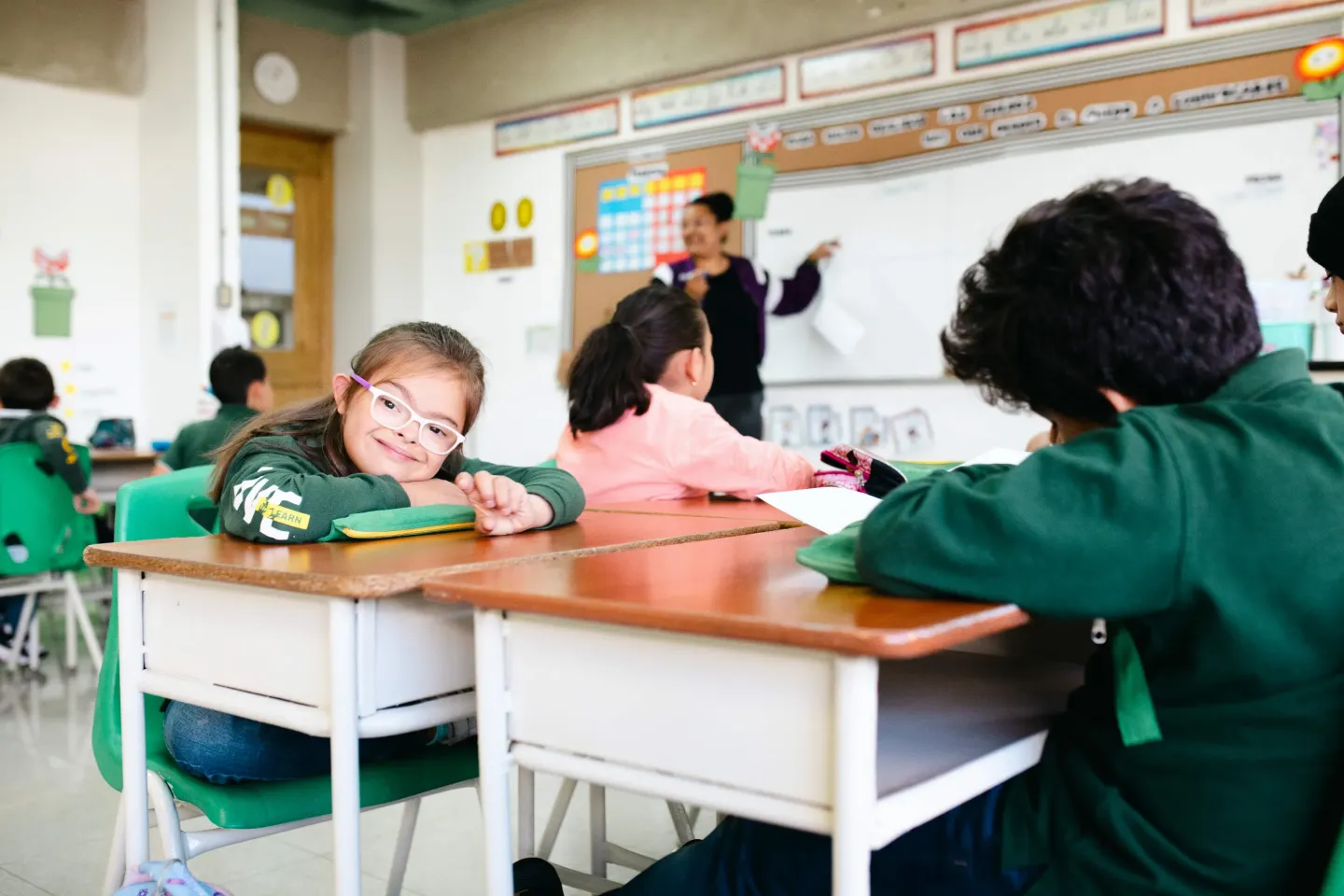 A girl sitting at a school desk in a classroom stares at the camara, behind her we can see more students and a teacher in front of a blackboard.