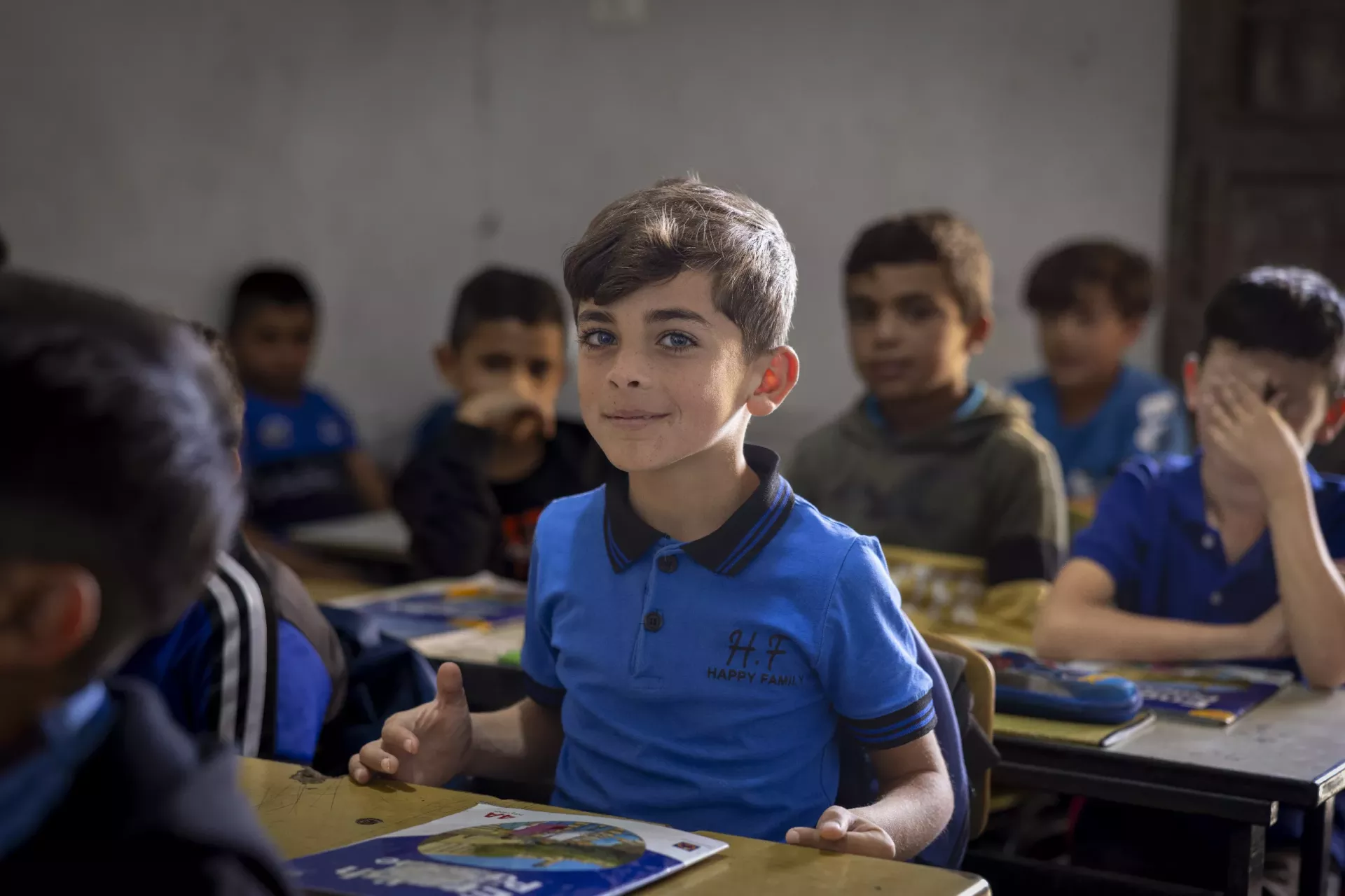 A boy sitting at his desk in a classroom. He is looking at the camera and smiling