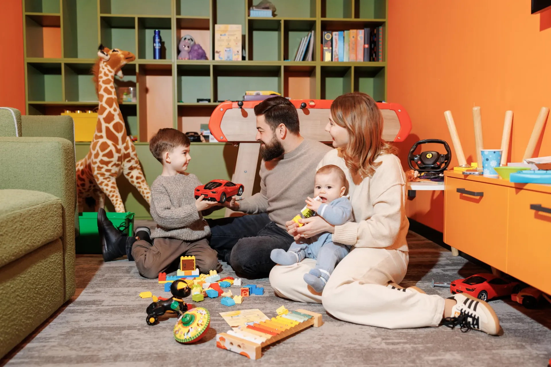 Parents sit on the floor with their small children surrounded by toys