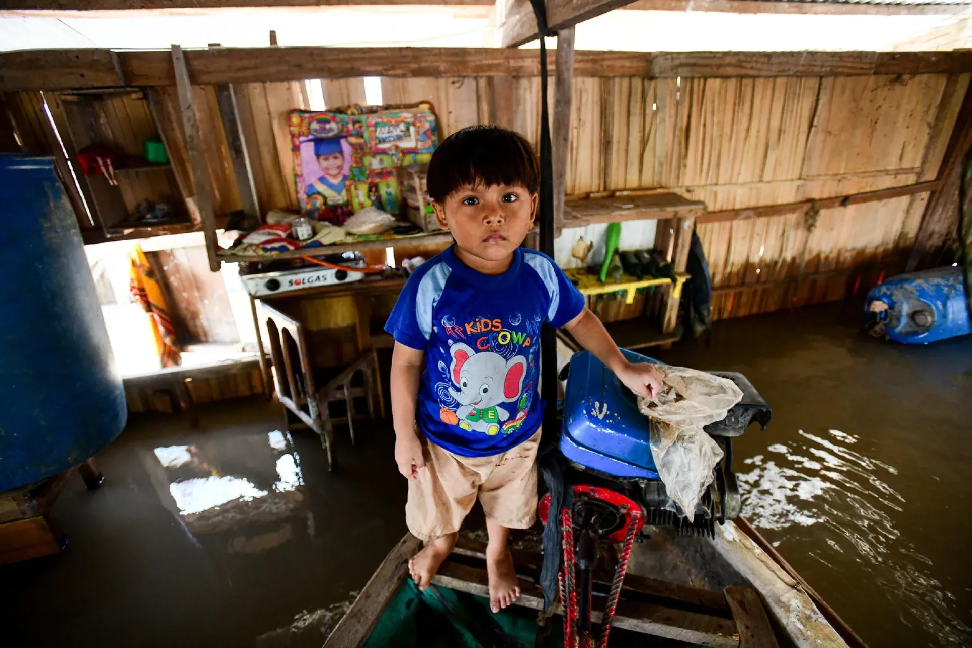 Young child stands on a makeshift boat indoors after heavy rains and flooding