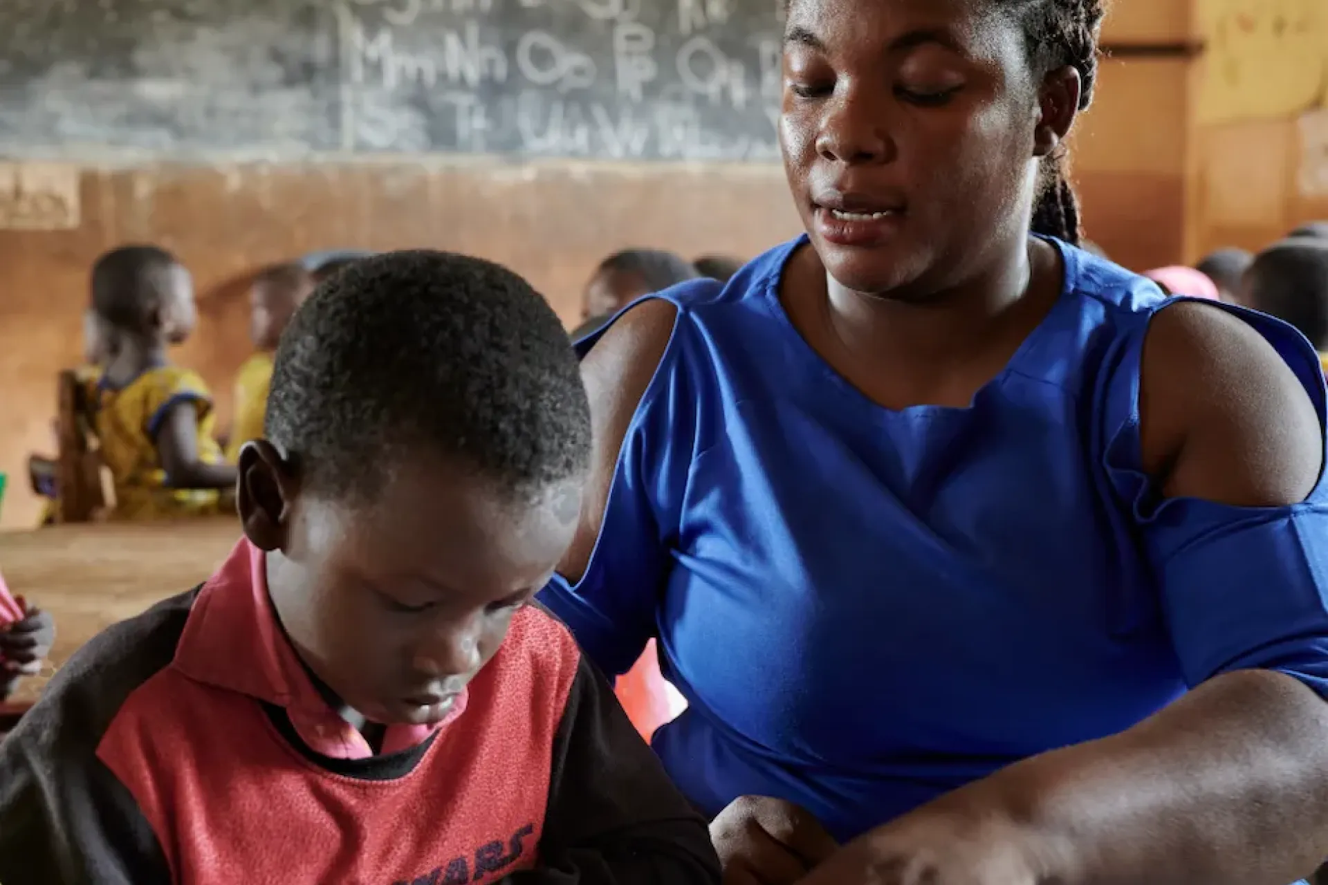 A teacher sits next to a child with a blackboard in the back, in a classroom in Ghana.
