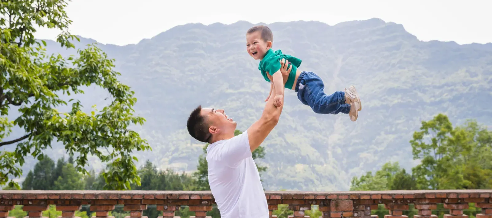 Adult lifting smiling child into the air on a terrace, mountains in background.