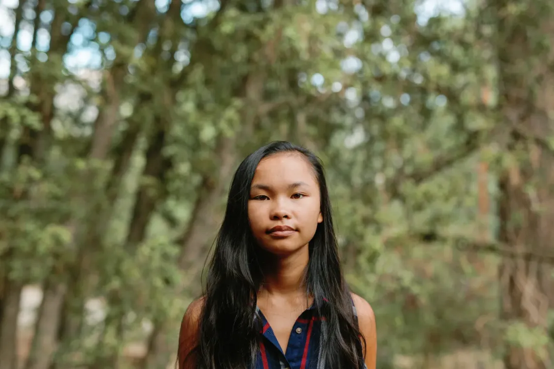 Teenager with long dark hair standing in front of a forest, looking straight into the camera