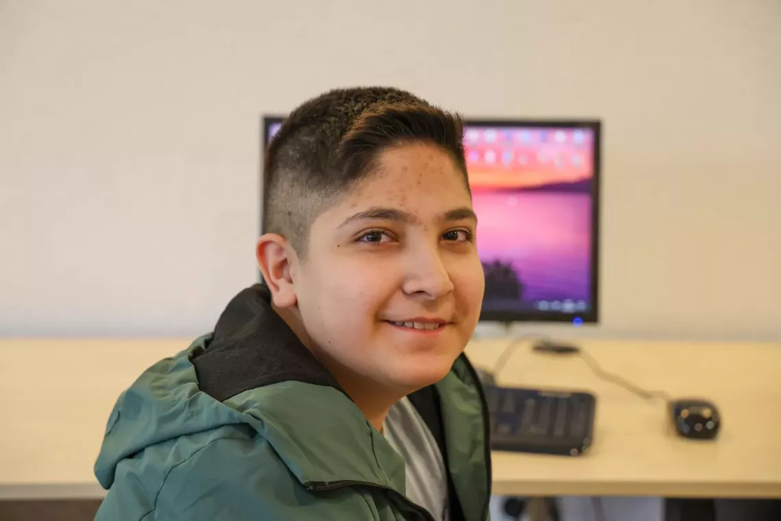 A young boy facing the camera and smiling, a computer can be seen on the desk behind him