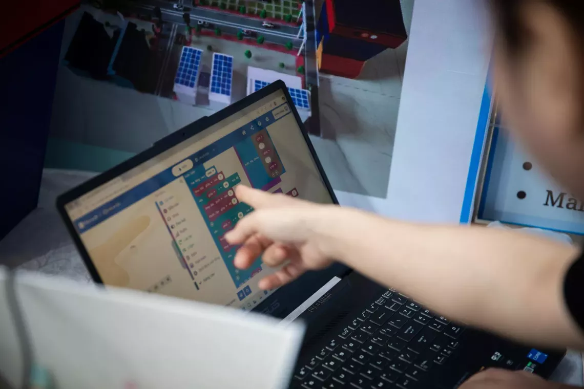 A child, out of focus and in the foreground, points to the screen of a laptop computer