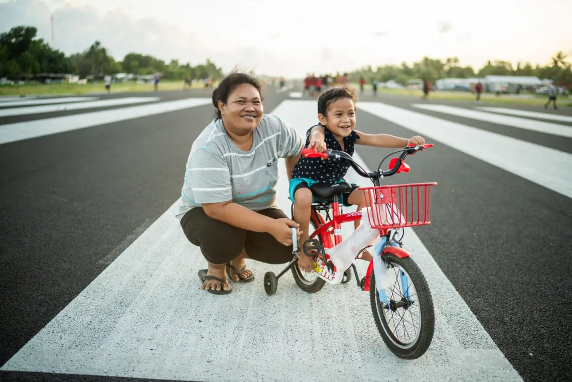 Mother squatting down beside her young child who rides a bike with training wheels