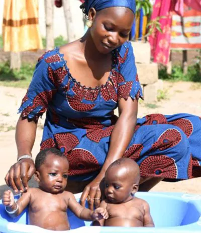 A woman gives two baby boys a bath in a blue plastic tub outside 