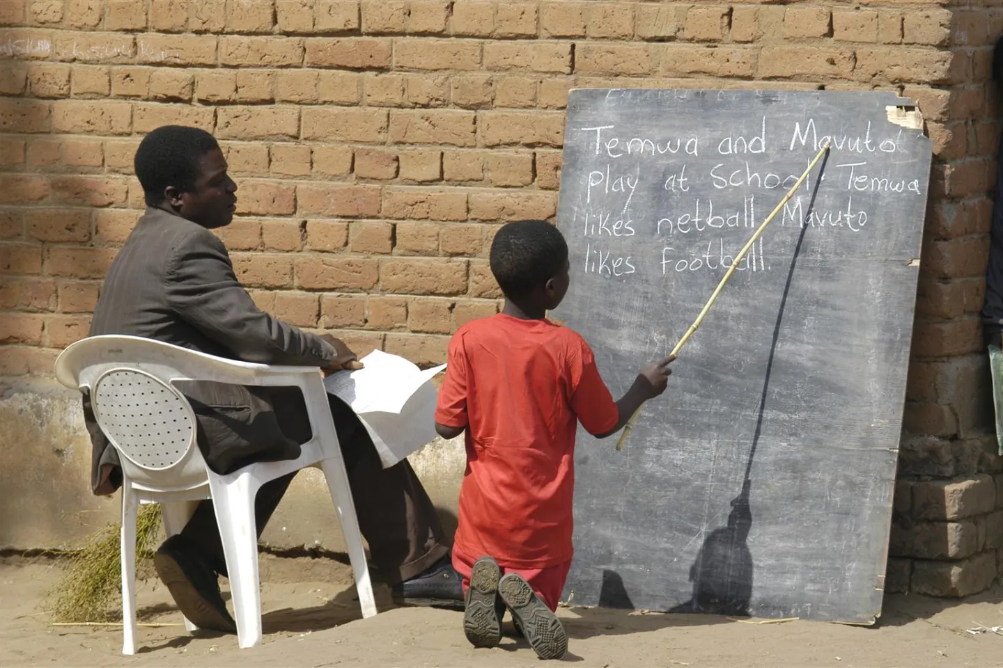 With his teacher seated beside him, a boy takes an English reading test kneeling in front of a blackboard that is propped against an outside wall at Chadza Primary School in a rural district near Lilongwe.