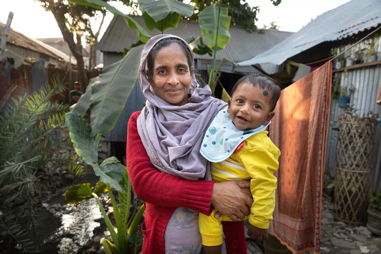 A woman holds a smiling baby outside an informal settlement with green leaves and corrugated metal roofs