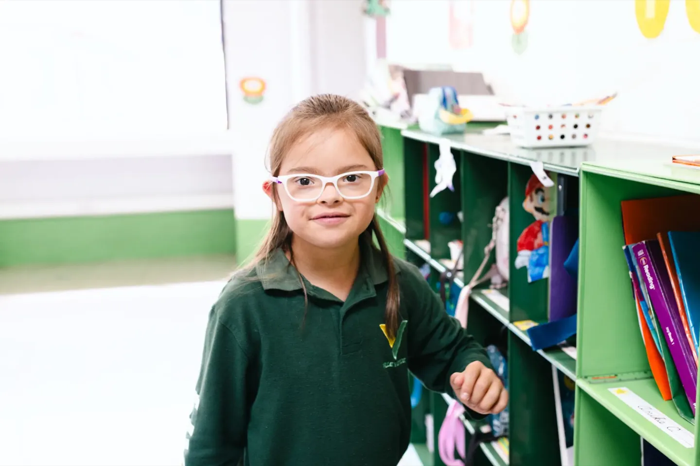 A young girl in glasses and a green school uniform smiles beside classroom shelves in Guatemala City.