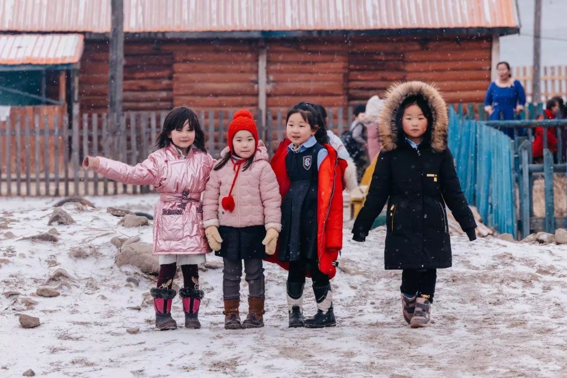 A group of four girls wrapped up warm in the snow and ice outside a wooden school structure.