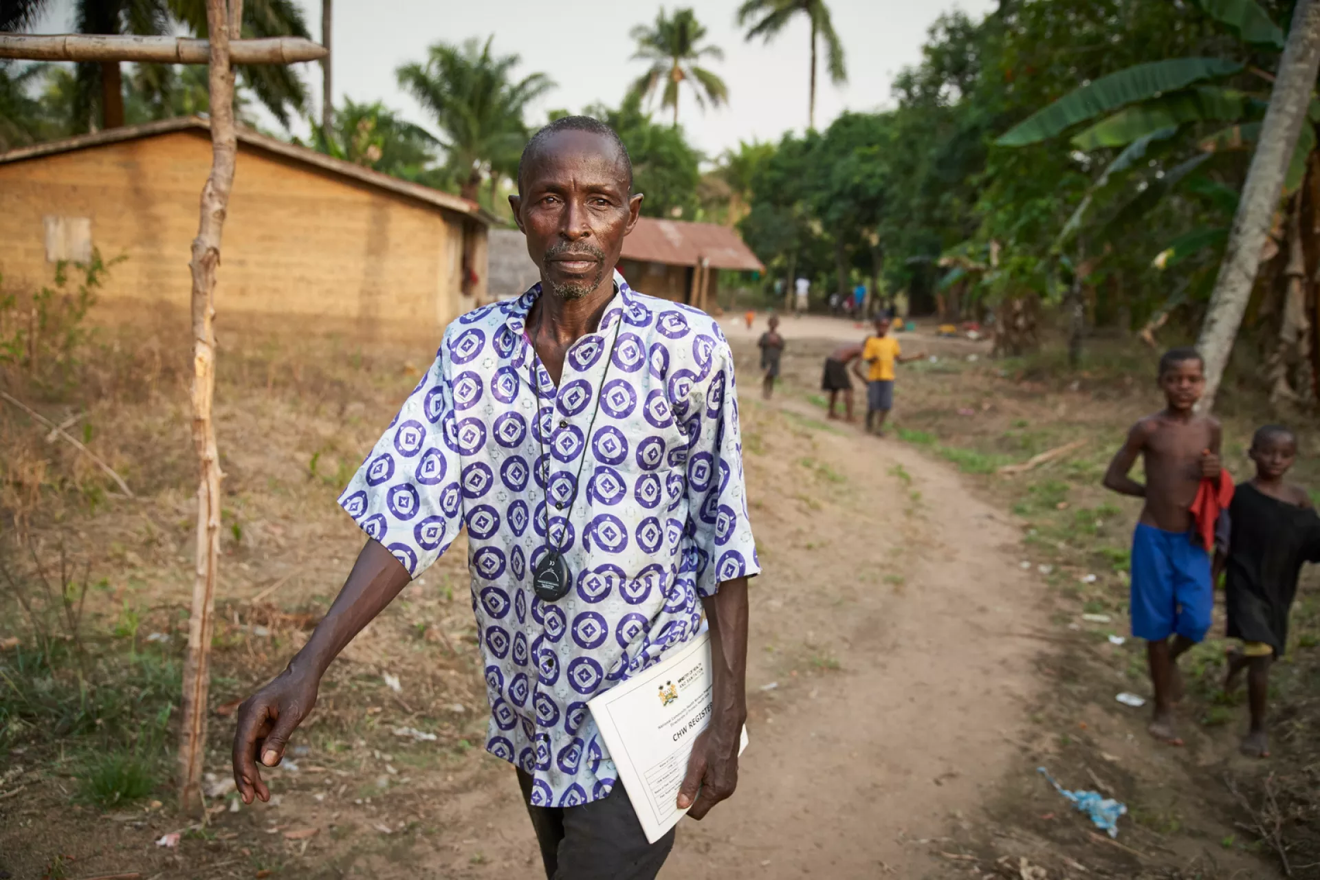 Community Health Worker (CHW) Brima Bangura, carrying health papers, walks through Maforay Village in Safroko Limba Chiefdom, Bombali District. Two boys are visible behind him. Mr. Bangura became a CHW in 2012 to help bring basic maternal and child health services closer to his village.