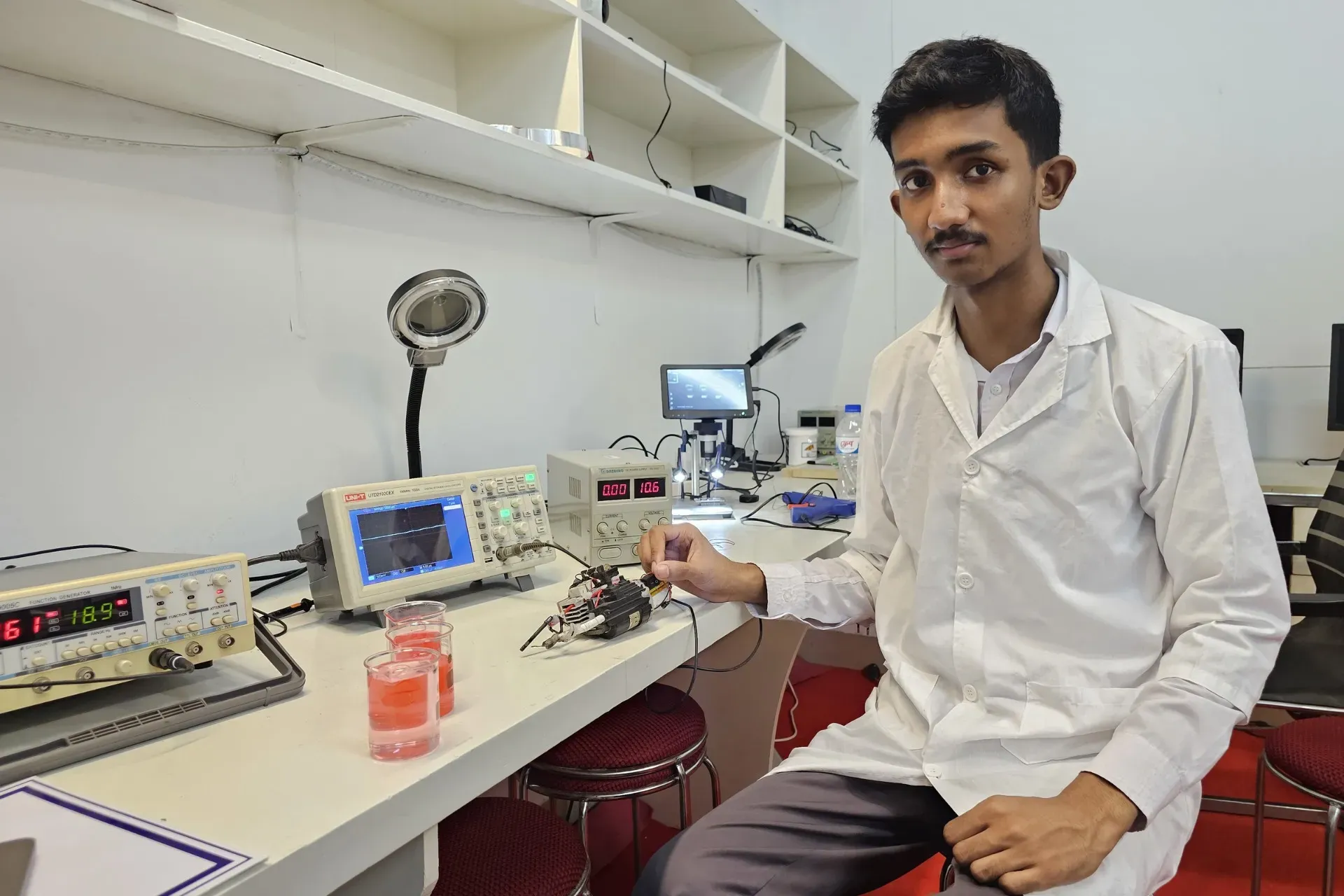 A young man in a lab coat sitting at a desk with electronic and scientific equipment.