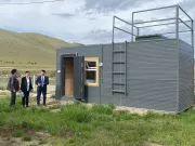 A grey cold sanitation waste water treatment container with a ladder on the outside leading to a platform on top with railings. The door is open, showing a wooden inside. One woman and two men stand outside in a setting with soft rolling hills and lush green grass.