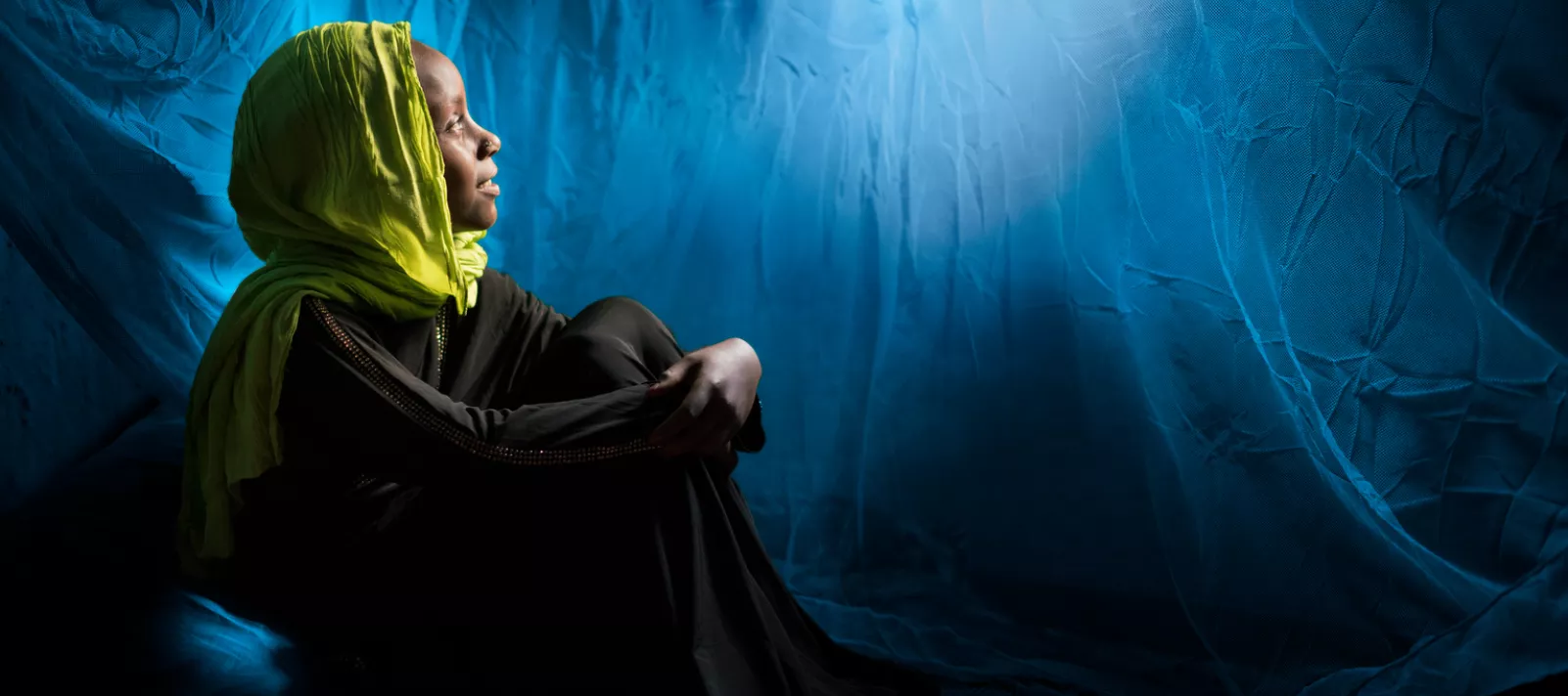 A young girl wearing a colourful headscarf sits under a mosquito net and looks up at light filtering in 