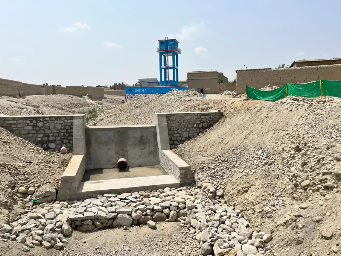 A concrete engineered structure leading into a dry riverbed with a UNICEF branded water tower in the background.