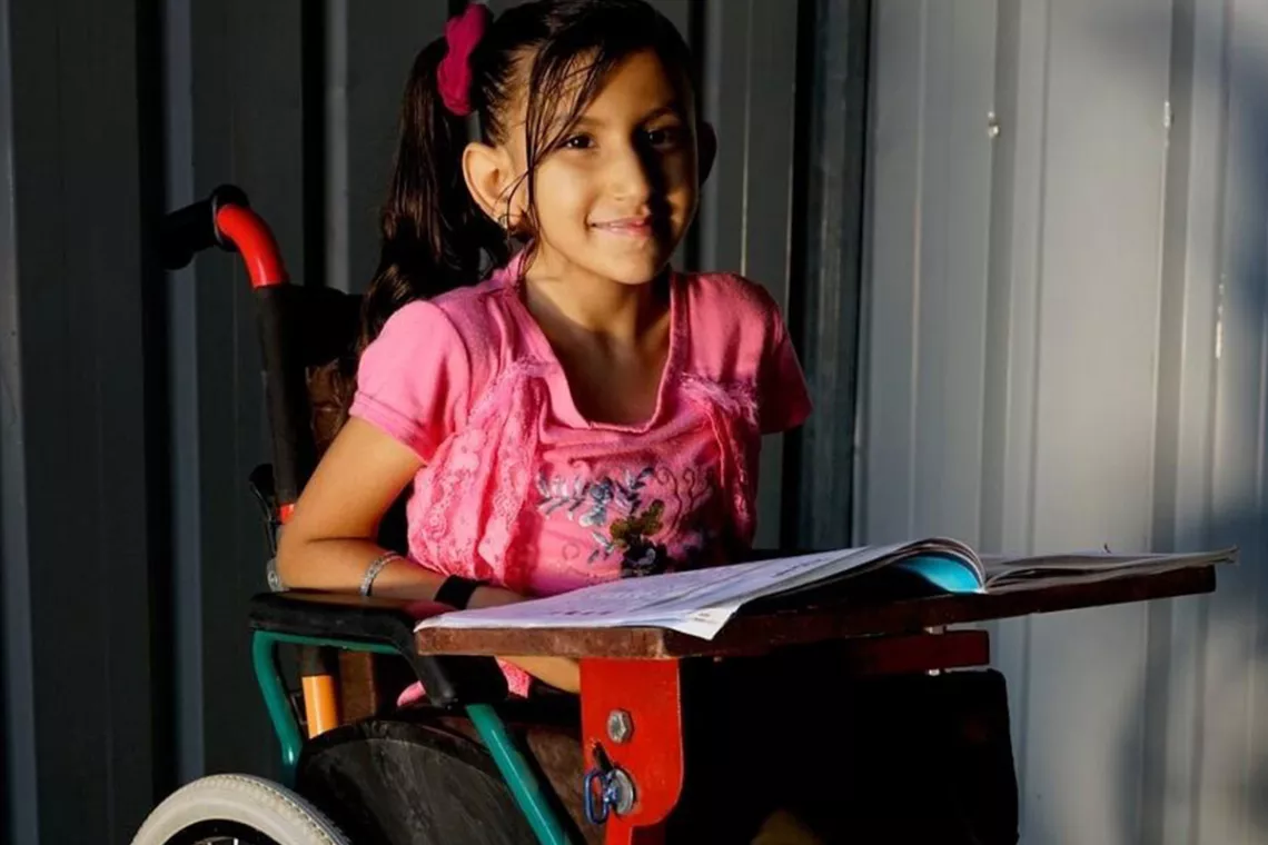 A girl in a wheelchair takes a break from reading a book to smile for the camera