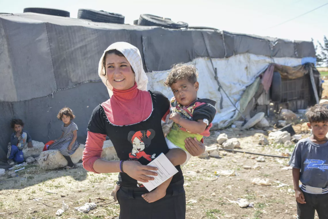 A mother holding her child and the vaccination card in an informal settlement of displaced families on Hama Highway, Homs Governorate, Syria