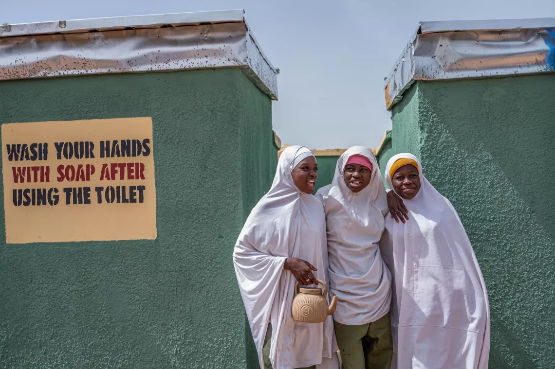 Three girls smile outside a block of toilets.