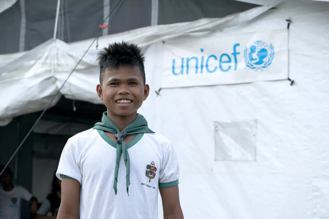 A young boy standing in front of a UNICEF tent smiles for the camera