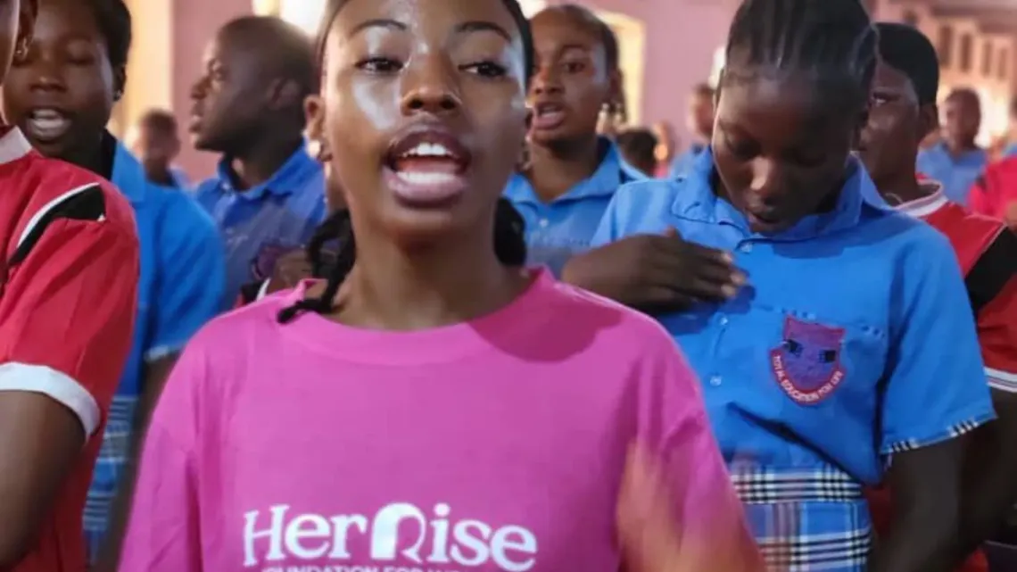 A girl wearing a pink t-shirt with the words 'Her Rise' speaks at an event