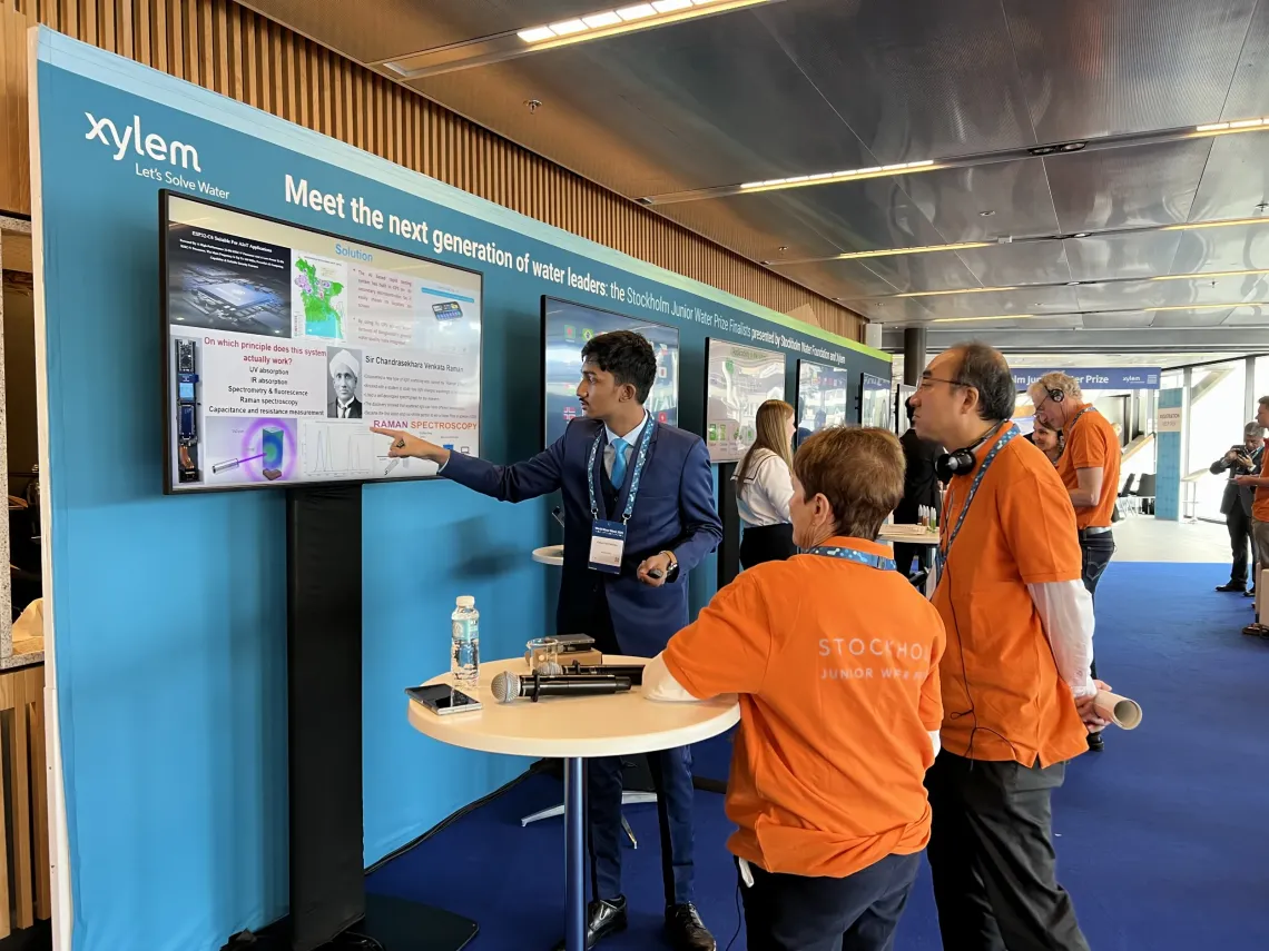 A young man in a suit points at a screen at an event as a woman and a man with orange t-shirts attentively watch.