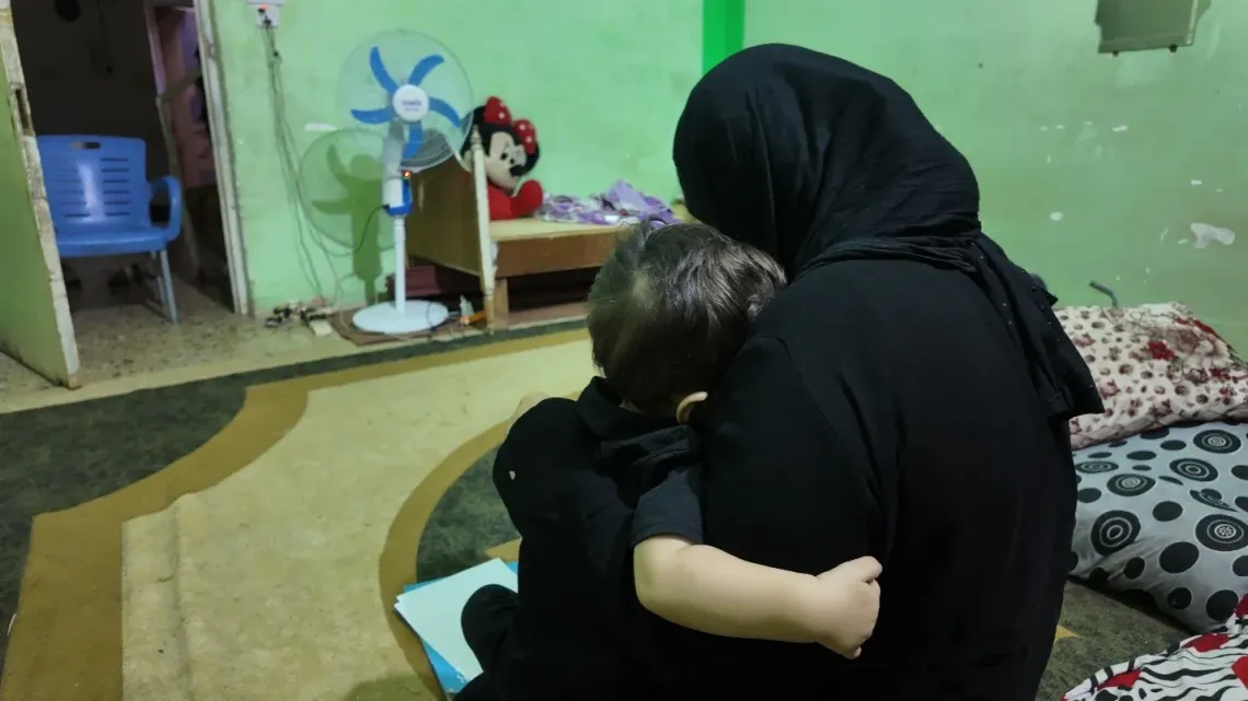 Woman in a black headscarf hugging a small child in a room with a fan and toys.