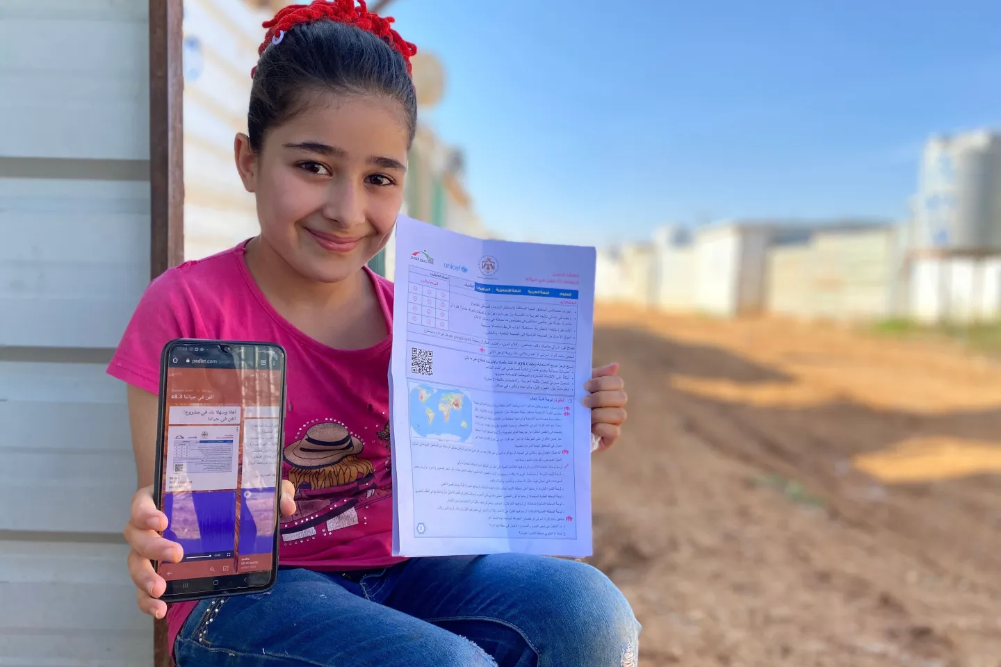 photo of a girl sitting outside in a refugee camp setting holds a mobile phone and sheet of paper up 