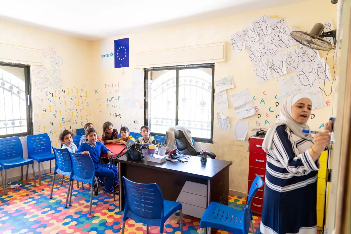 Children sit at a classroom table as a teacher writes on the whiteboard