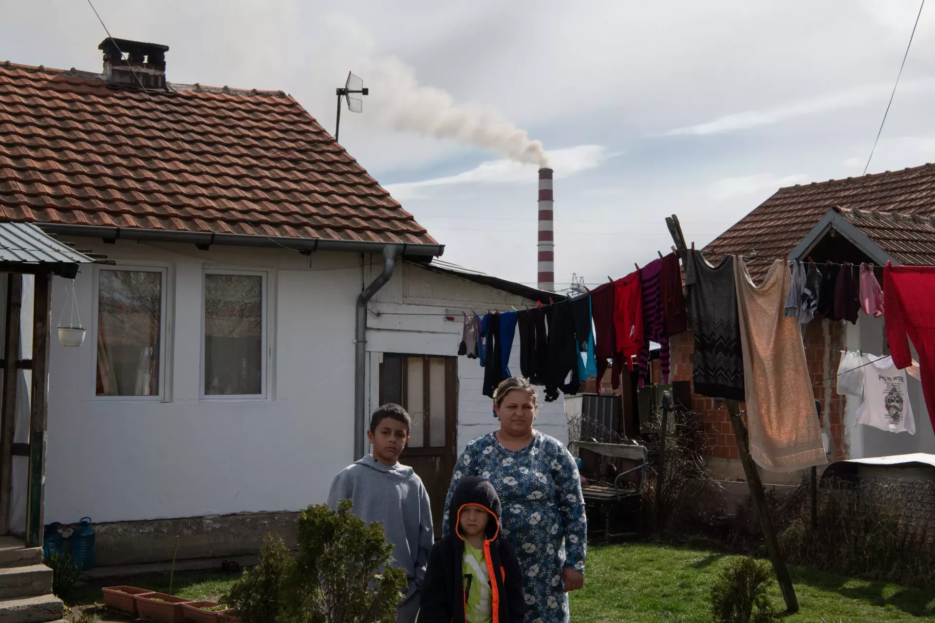 Nexhare stands with her two sons in the garden of their home in Obiliq, Kosovo. Power Plant A, visible in the background, is polluting the air, making her children sick, March 2024.