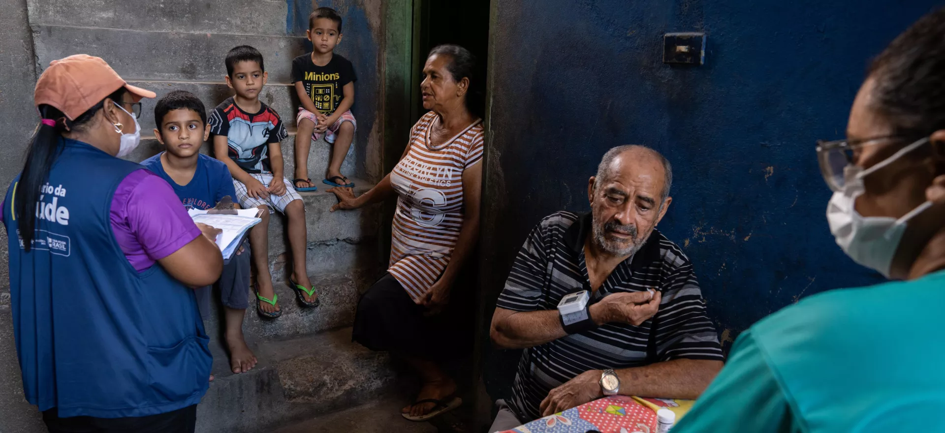 Community Health Agent talks to an older woman at her house, with her grandsons sitting on the stairs listening.