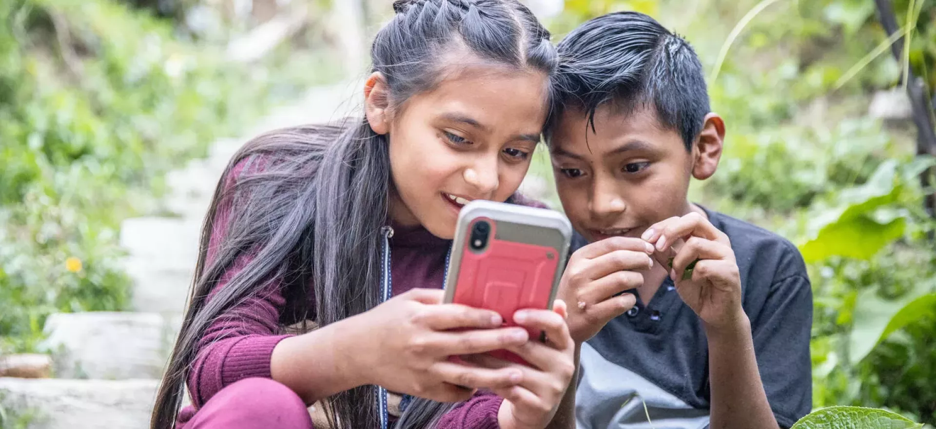 Una ni&ntilde;a y un ni&ntilde;o sentados en medio de un jard&iacute;n sosteniendo un tel&eacute;fono m&oacute;vil en sus manos