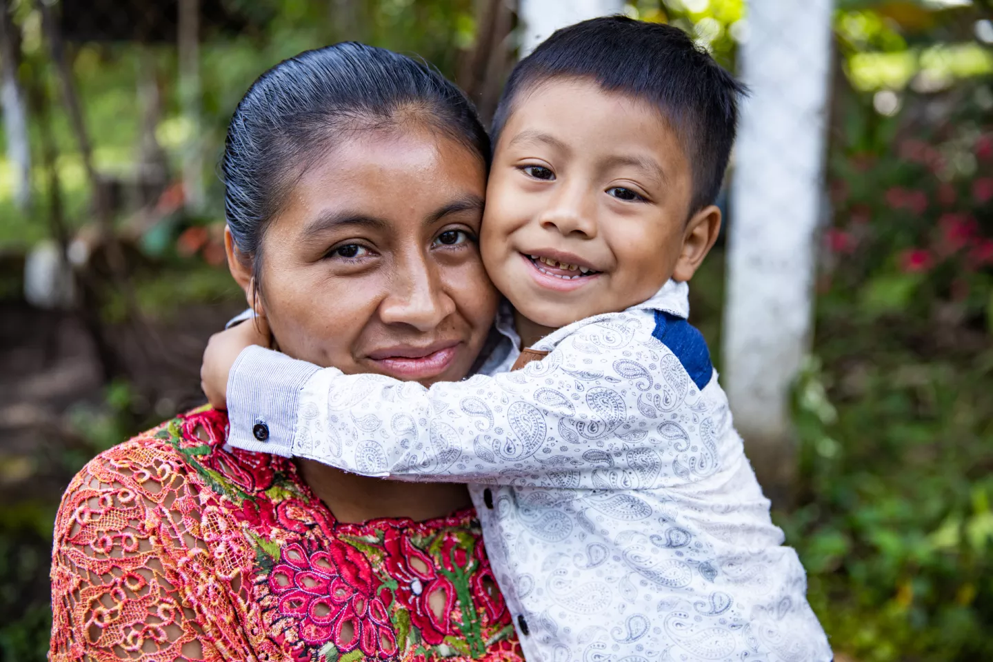 A little boy hugs his mother. They both look at the camera and smile. 