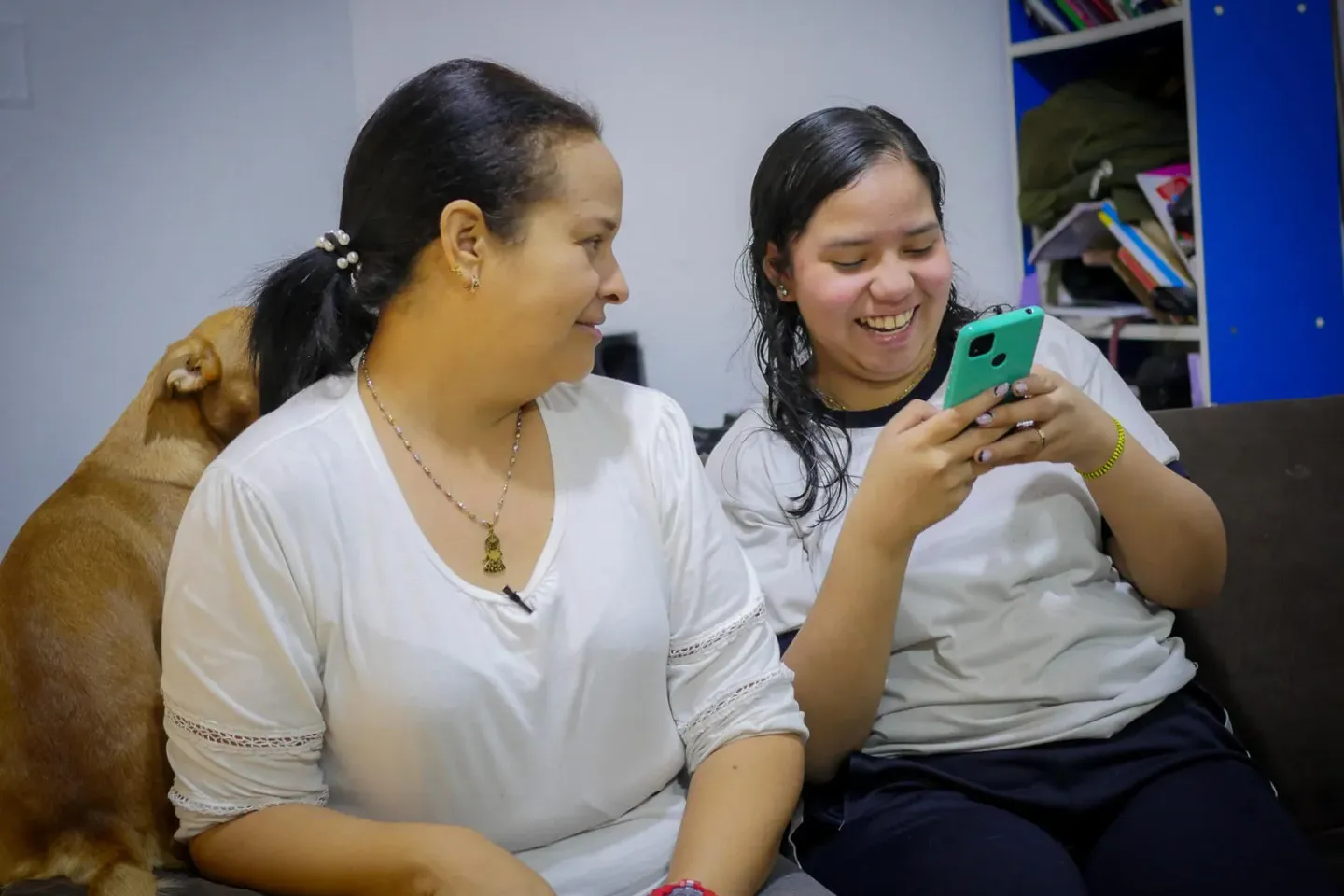 Una mujer y una adolescente sentadas en un sofa, la mujer adulta mira a la adolescente mientras ella usa un tel&eacute;fono celular.