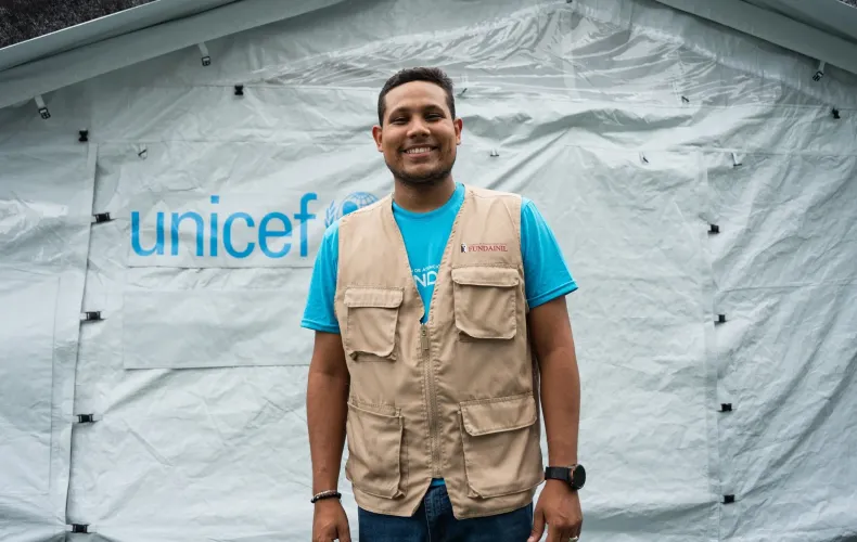 Un hombre de pie, sonriendo, frente a una carpa en un campamento transitorio