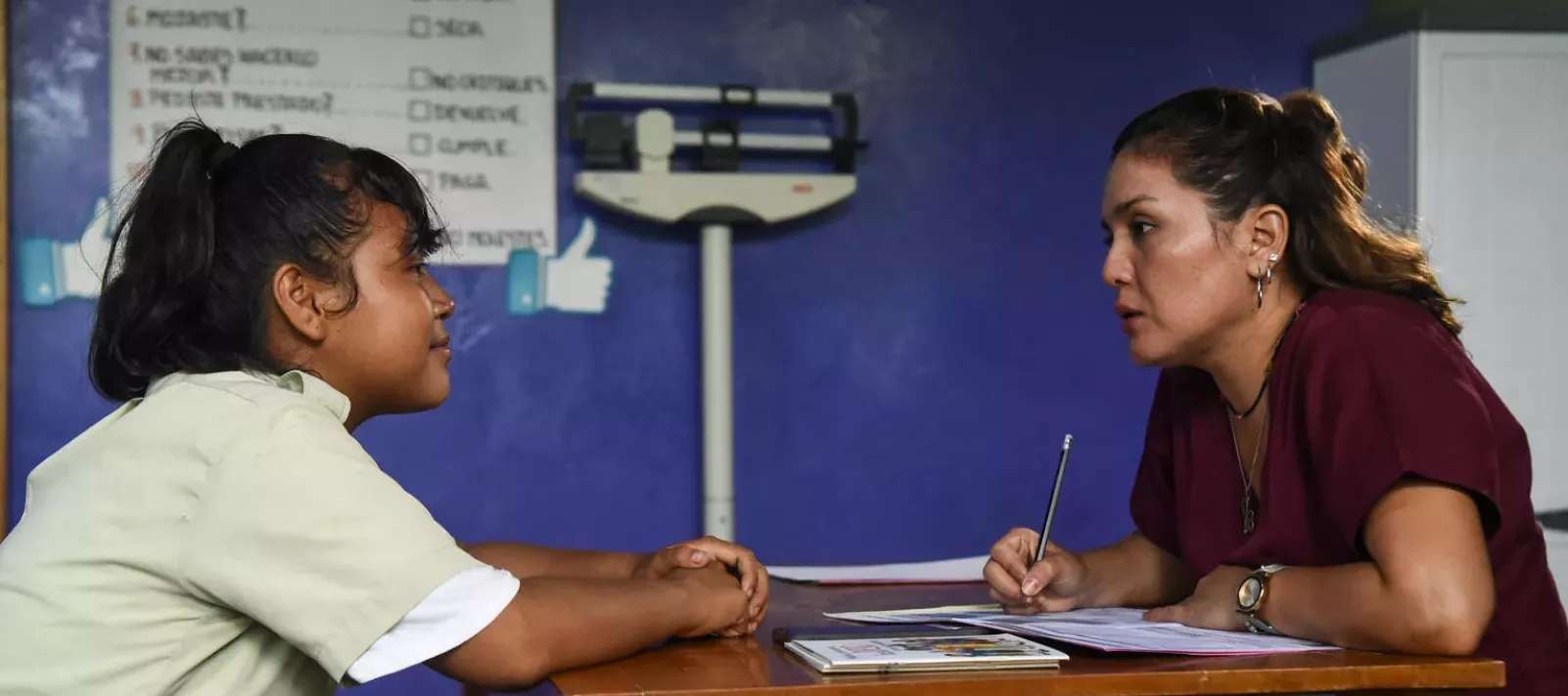 Una adolescente, sentada de un lado de un escritorio oye mientras una doctora le habla