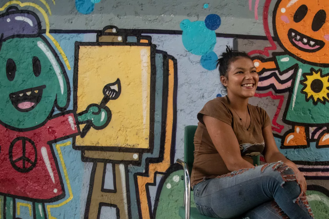 Una joven sonriendo y tiene como fondo un mural.