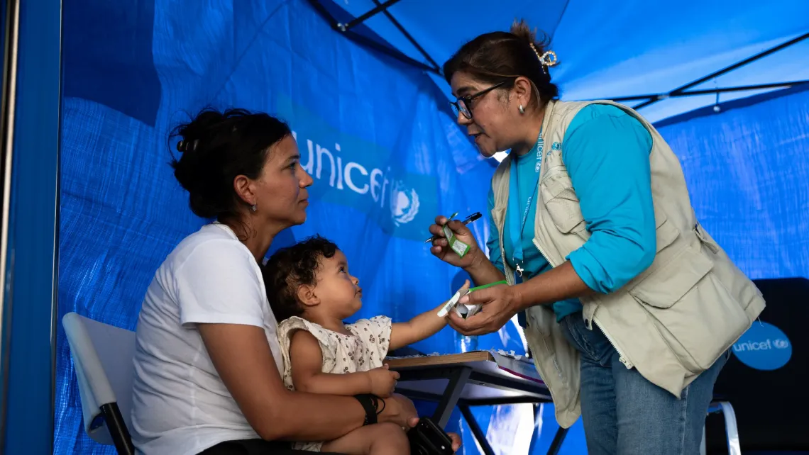 Una madre sostiene a su bebe mientras recibe informaci&oacute;n de una trabajadora de UNICEF, dentro de una carpa