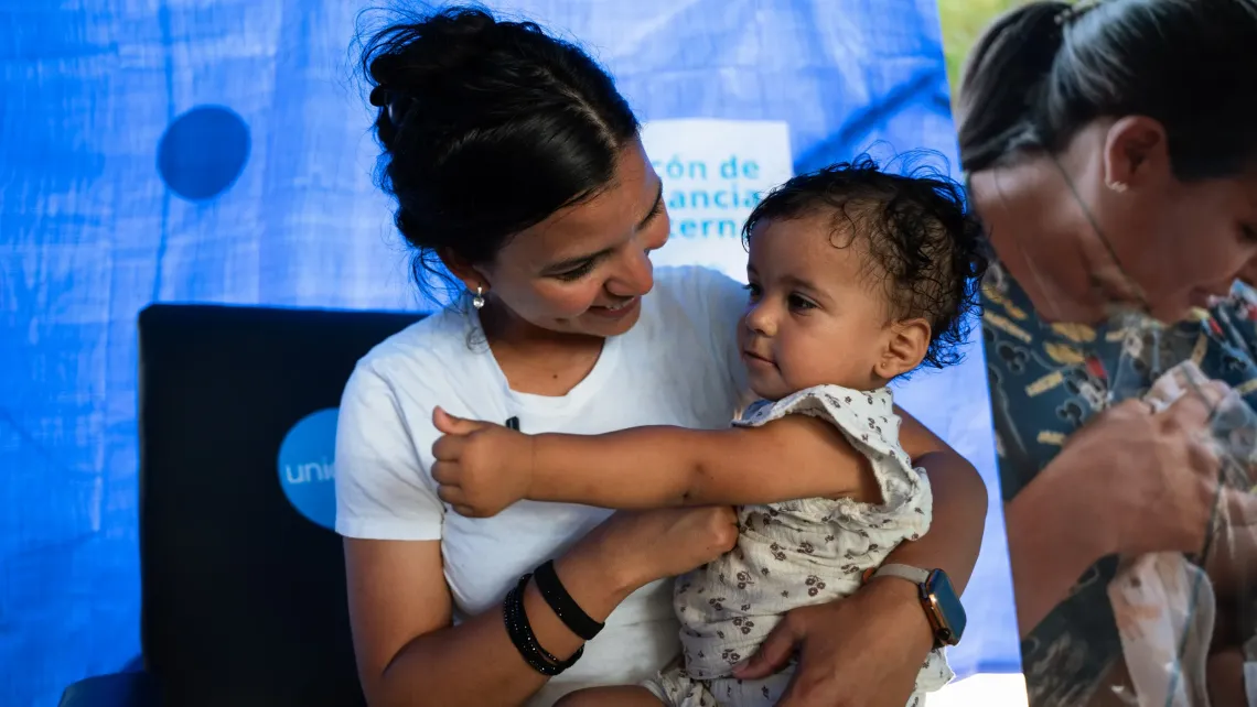 Una madre sostiene a su bebe mientras ambas sonr&iacute;en, dentro de una carpa de UNICEF