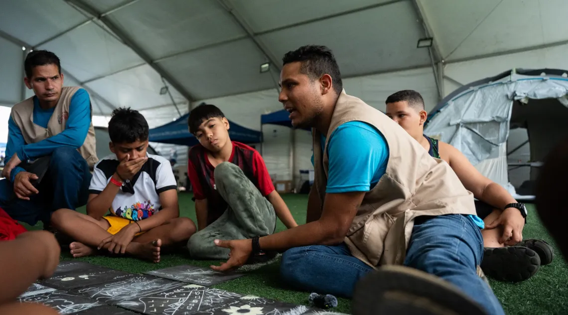 Un hombre sentado habla mientras un grupo de ni&ntilde;os alrededor le presta atenci&oacute;n.