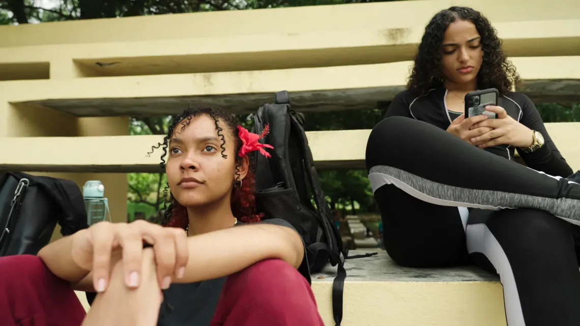 Dos adolescentes sentadas, una mirando la pantalla de un tel&eacute;fono, la otra mirando hacia adelante
