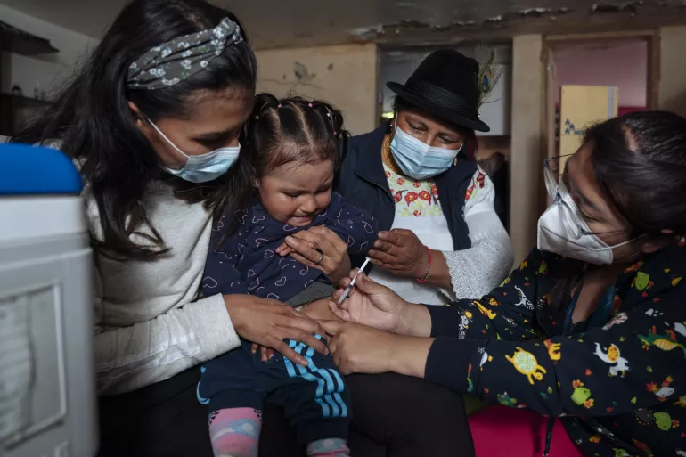 Una ni&ntilde;a ecuatoriana de 2 a&ntilde;os, recibe una vacuna en su casa mientras su madre, la carga.
